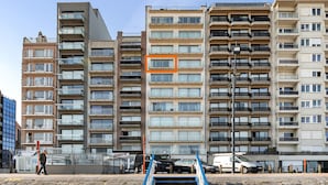 Un immeuble résidentiel à plusieurs étages, doté de balcons, avec un rectangle orange bien visible qui délimite une partie du bâtiment, et une personne qui marche sur le trottoir.