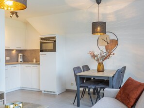 A modern kitchen with white cabinets, a built-in oven, and a coffee maker. There is a dining area with a table, chairs, and a vase with dried flowers. - Apartment With Cosy Sunny Terrace (Middelkerke)