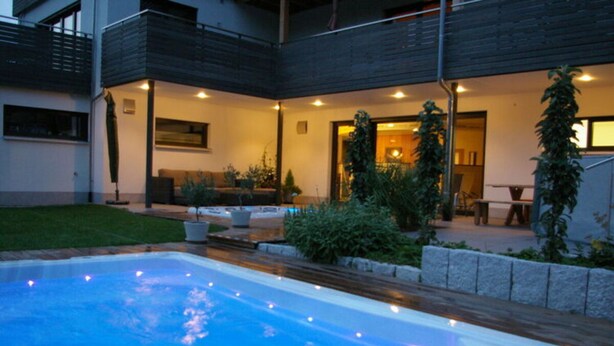 Uma área moderna com piscina ao ar livre e hidromassagem, rodeada por um deck de madeira e plantas em vasos.