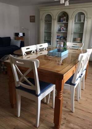 A dining area with a glass-top table surrounded by white chairs with blue cushions. There is a cabinet with glass doors in the background. - Maisonette to Gottorf and Schlei (Schleswig)