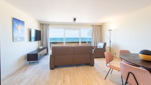 Een ruime woonkamer met een grote bank, een eettafel en een televisie. Achter de glazen deuren is een balkon met uitzicht op de oceaan zichtbaar.