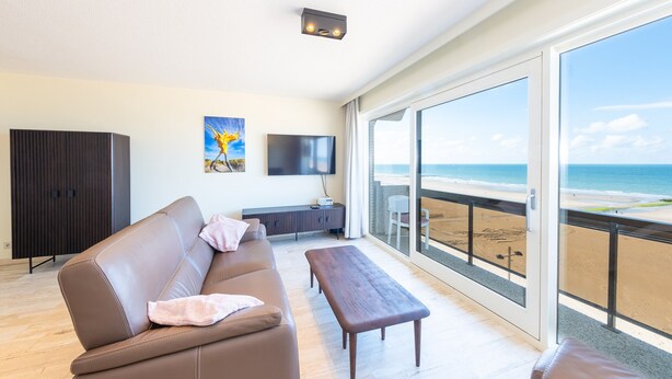 Een moderne woonkamer met een bank, een salontafel en een televisie. Er is een groot raam met uitzicht op het strand.