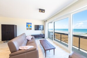 Een moderne woonkamer met een bank, een salontafel en een televisie. Er is een groot raam met uitzicht op het strand.