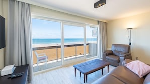 Een hotelkamer met een grote schuifglazen deur die uitkomt op een balkon met uitzicht op het strand. Er is een bruine leren bank, een klein houten tafeltje en een vloerlamp.