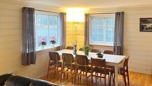 Un comedor con una mesa dispuesta para seis comensales, flanqueado por ventanas con cortinas, una lámpara y un cuadro en la pared.
