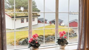 Una ventana con dos macetas blancas de flores rosadas en el alféizar, con vista a una zona costera con casas y una extensión de agua.
