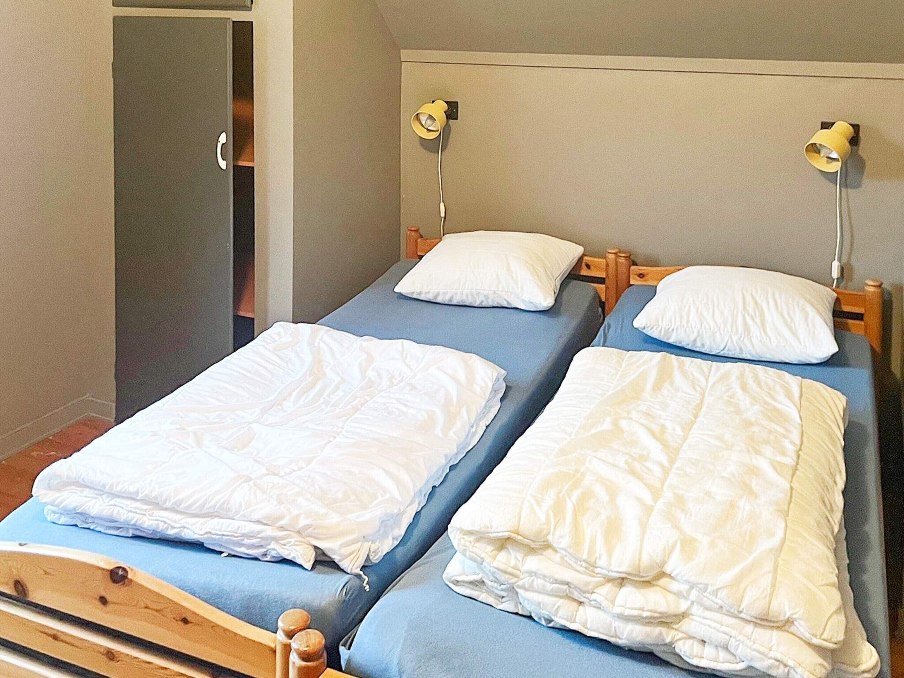 A room with two single beds, each with a wooden headboard and a blue mattress cover. There are two bedside lamps on each bed.