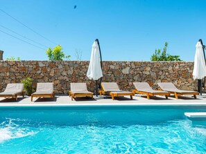 A poolside area with lounge chairs and umbrellas, a stone wall, and a clear blue pool. - With Pool, Jacuzzi and Sauna (Privlaka)