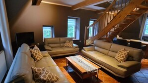 Ein geräumiges Wohnzimmer mit zwei Sofas, einem Couchtisch und einem Fernseher. Im Hintergrund ist eine Holztreppe zu sehen.