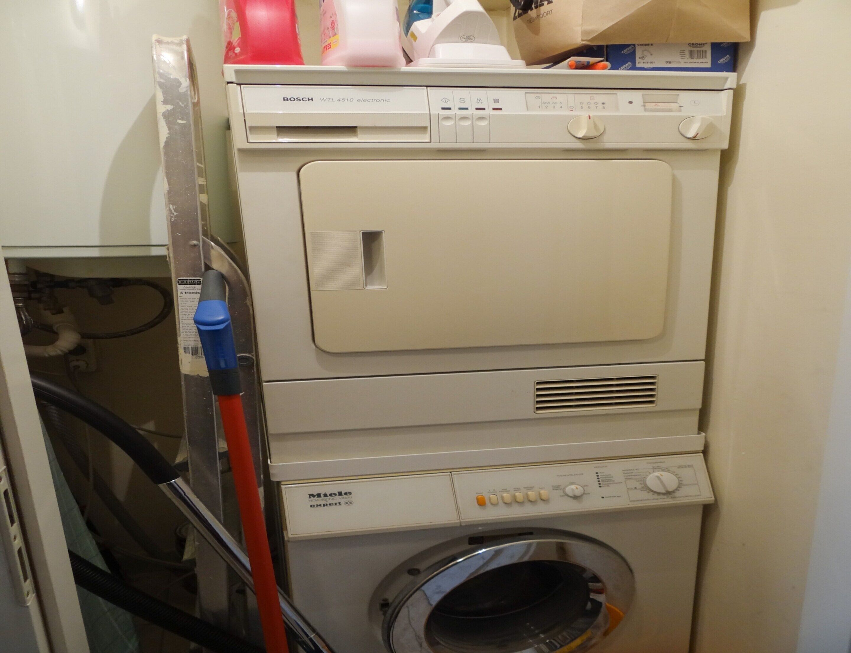 A stacked washer and dryer unit with a control panel, a hose, and a paper bag on top.