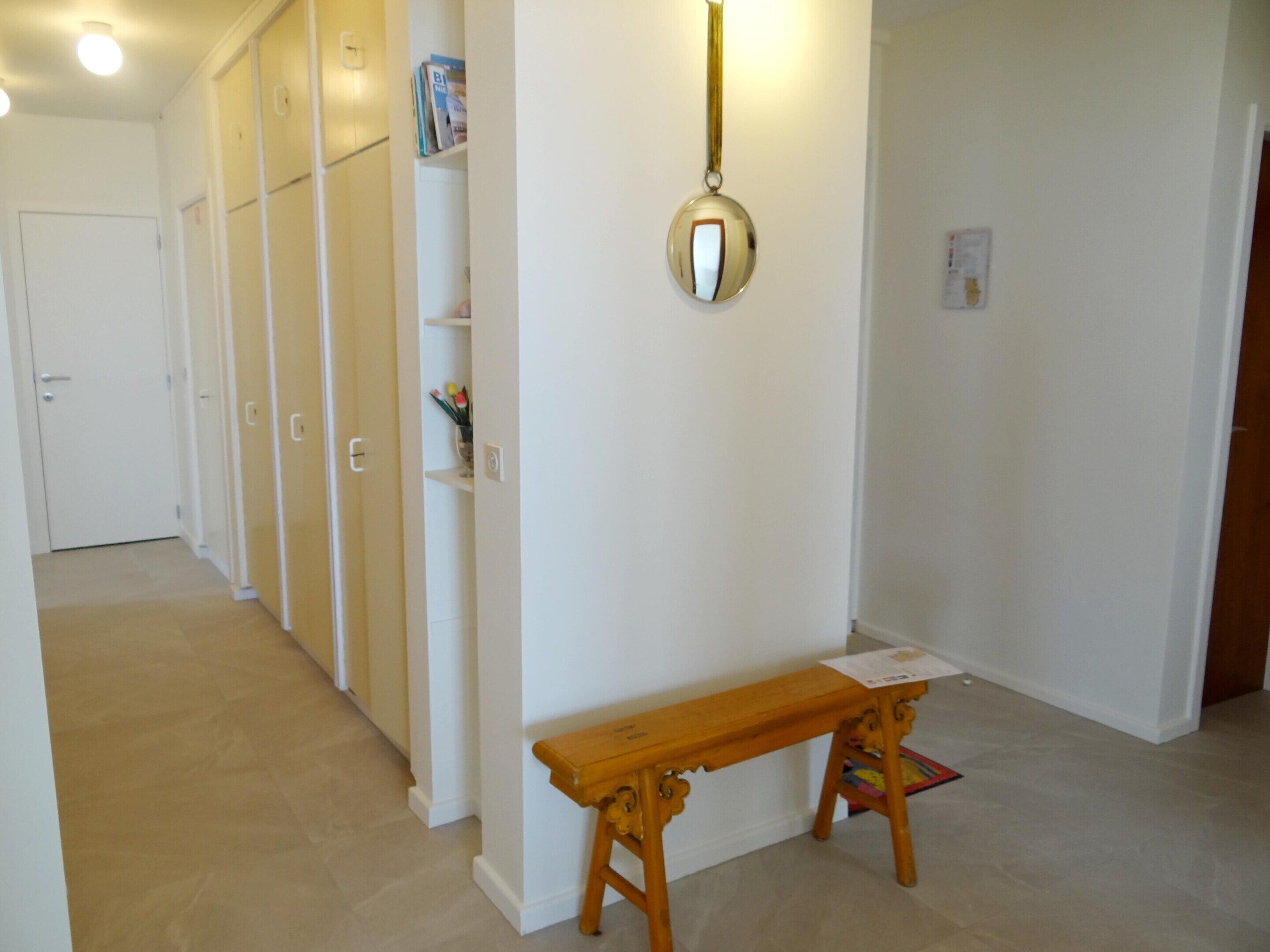 Un couloir avec un banc en bois, un miroir au mur et une porte au fond.