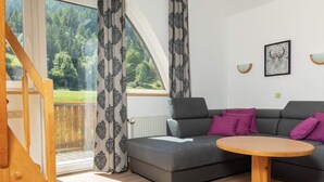 A modern living room with a grey sofa, purple cushions, a wooden coffee table, and a view of a forest through large windows. - Apartment in the ski and Hiking Area in Mallnitz (Mallnitz)