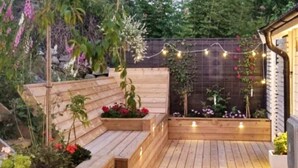 Une terrasse en bois dotée de bancs intégrés et de jardinières, entourée d’un jardin où poussent différentes plantes et fleurs.