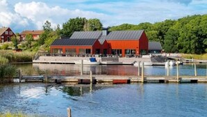 Een pand aan het water met een rode gevel, een steiger en veel groen eromheen.