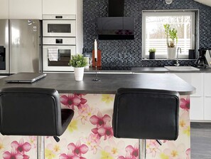 Una cocina moderna con una isla integrada, dos sillas negras, una planta en maceta y un ventanal con plantas afuera.
