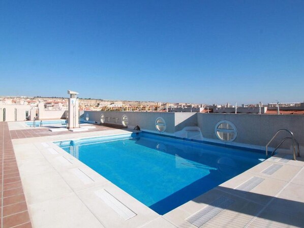 A rooftop swimming pool with clear blue water, surrounded by a tiled deck and a cityscape view in the background.