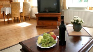 Un salon avec un meuble de rangement en bois pour l’équipement audiovisuel, une télévision, une table sur laquelle se trouve une assiette de fruits, une bouteille de vin et un vase rempli de fleurs.