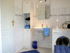 Une salle de bain équipée d’un lavabo, d’un miroir, d’un lave-linge et d’une poubelle bleue.