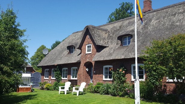 Une maison traditionnelle en briques, avec un toit de chaume, un mât sur lequel flotte un drapeau, et une pelouse agrémentée de deux chaises blanches.