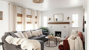A living room with a gray sectional sofa, a red armchair, a round coffee table, and a white fireplace. - Brick Haven House: King Suite, Near SUU, EV Charger, Parking, Nat’l Parks (Cedar City)