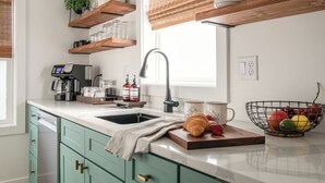 Una cocina con encimera blanca, un lavabo y una cafetera. Hay estantes de madera sobre la encimera con platos y una planta. Un frutero y un croissant están sobre la encimera.