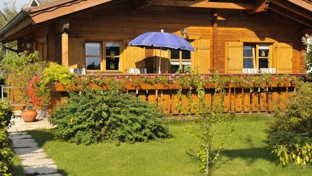 Una cabaña de madera con techo inclinado, un balcón con sombrilla azul y un jardín con diversas plantas y un sendero de piedra.