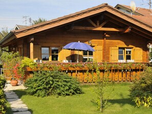 Eine Holzhütte mit geneigtem Dach, einem Balkon mit blauem Sonnenschirm und einem Garten mit verschiedenen Pflanzen und einem Kiesweg.