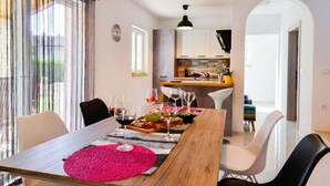 A dining area with a wooden table, white chairs, and a pink placemat. There is a kitchen in the background with white cabinets and a hanging light fixture. - Villa Stella Comfortable Holiday Residence (Umag)