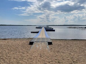 A sandy beach with a long wooden dock extending into calm water, surrounded by a clear sky.