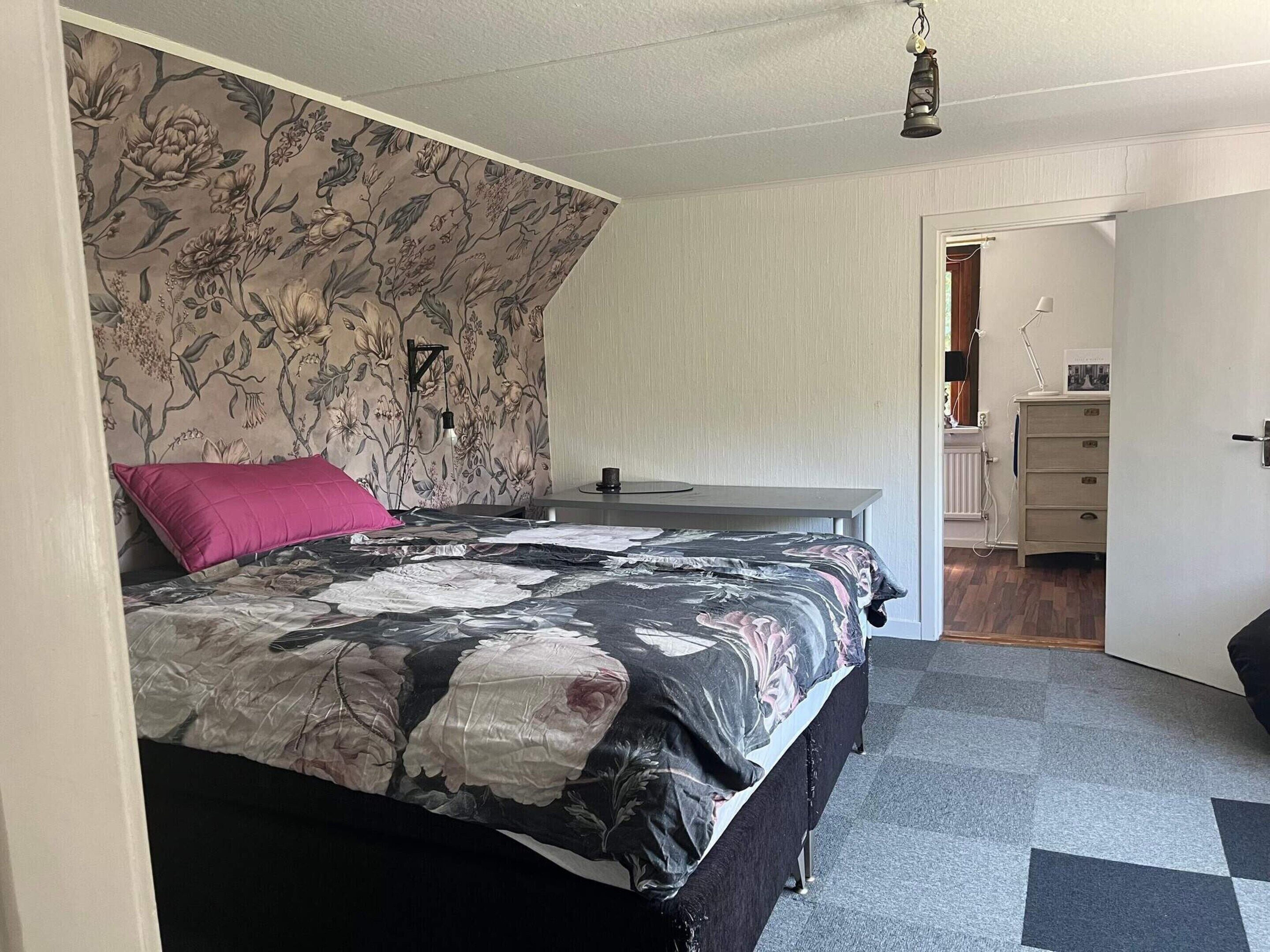 A bedroom with a floral-patterned headboard, a bed with a black and white floral bedspread, a pink pillow, and a bedside table with a lamp.