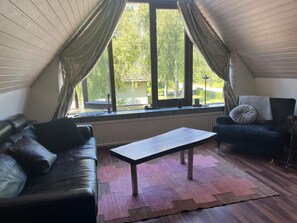 A room with a sloped ceiling, a black leather sofa, a dark armchair, a wooden coffee table, and a view of greenery outside through large windows. - 5 Person Holiday Home in Loddekopinge (Loddekopinge)
