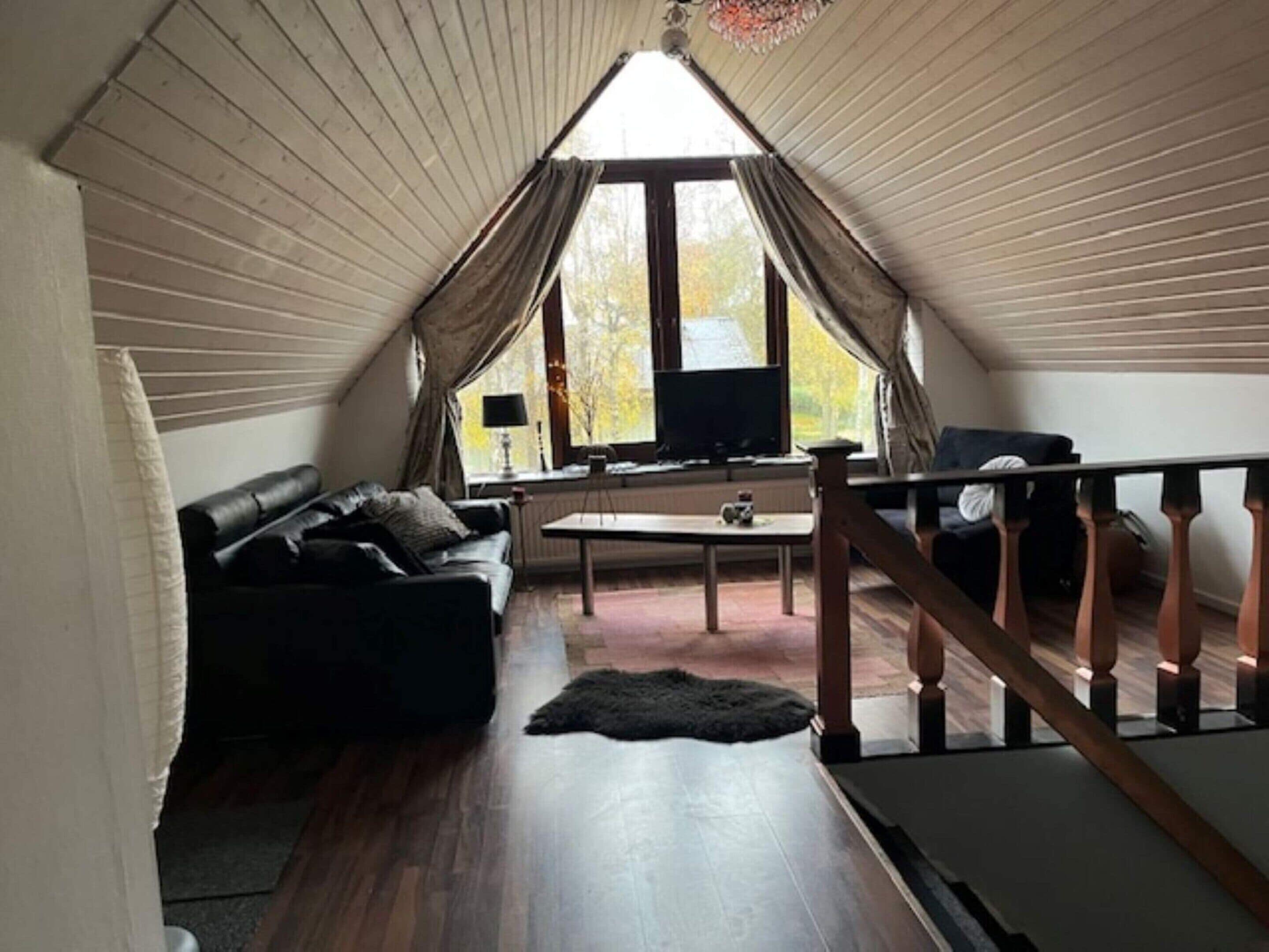 A cozy attic room with a wooden floor, a black leather sofa, a small table, and a TV.