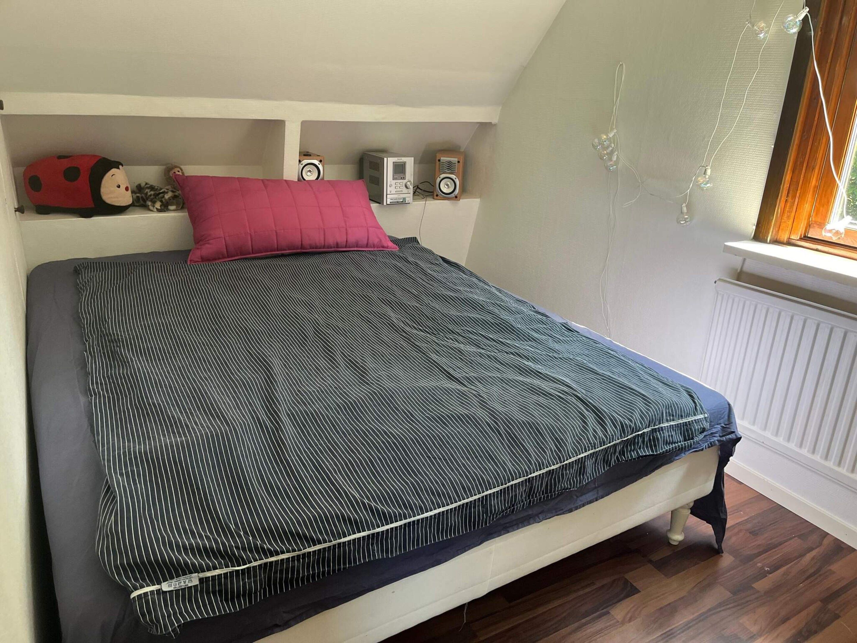 A bed with a striped bedspread, a red pillow, and a stuffed toy on the shelf above.