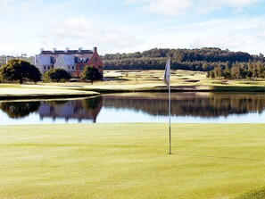 A golf course with a flagstick in the foreground, a pond reflecting the surroundings, and a building in the background.