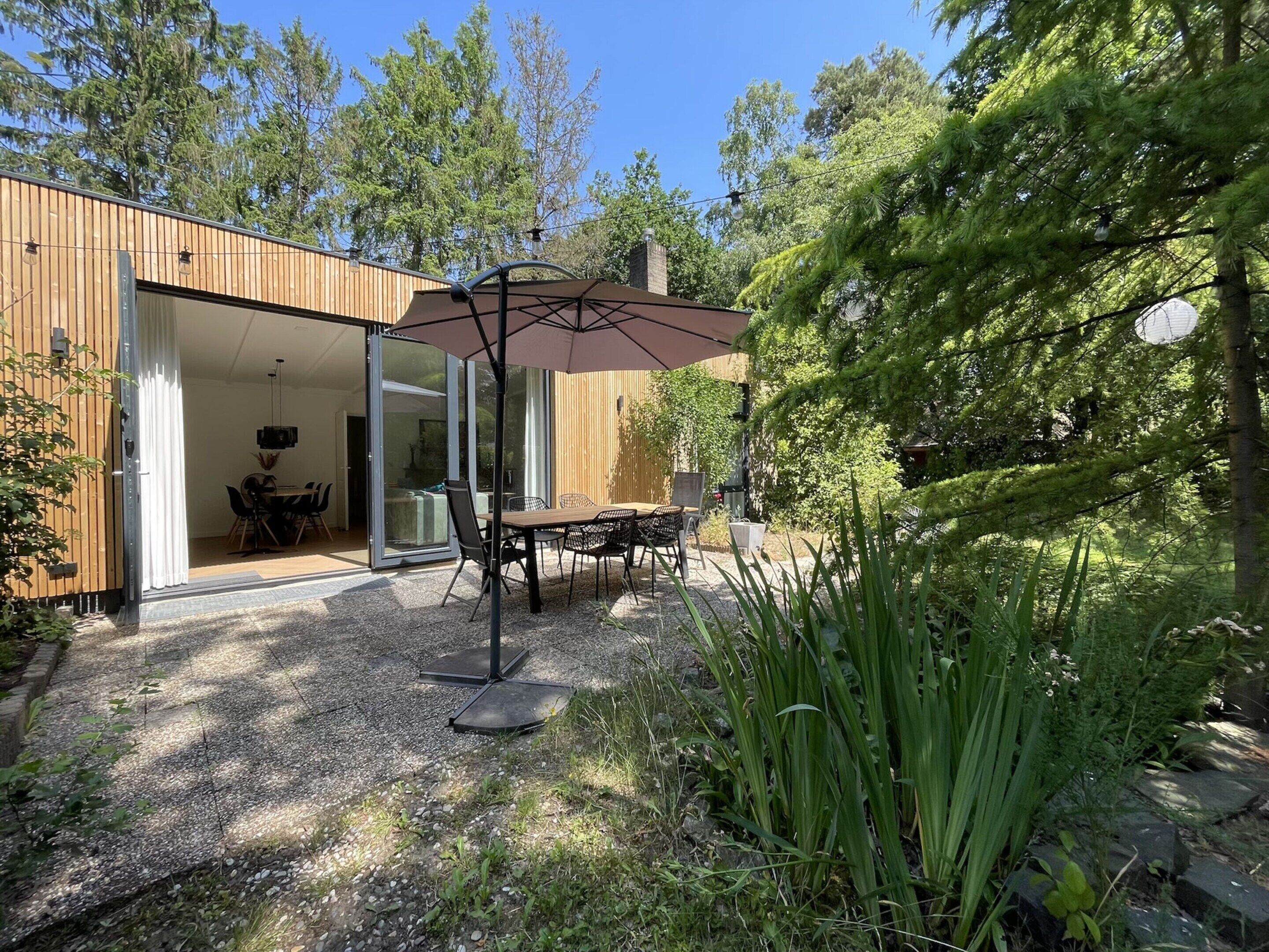 A modern house with a patio area featuring a table and chairs under a large umbrella, surrounded by natural vegetation.