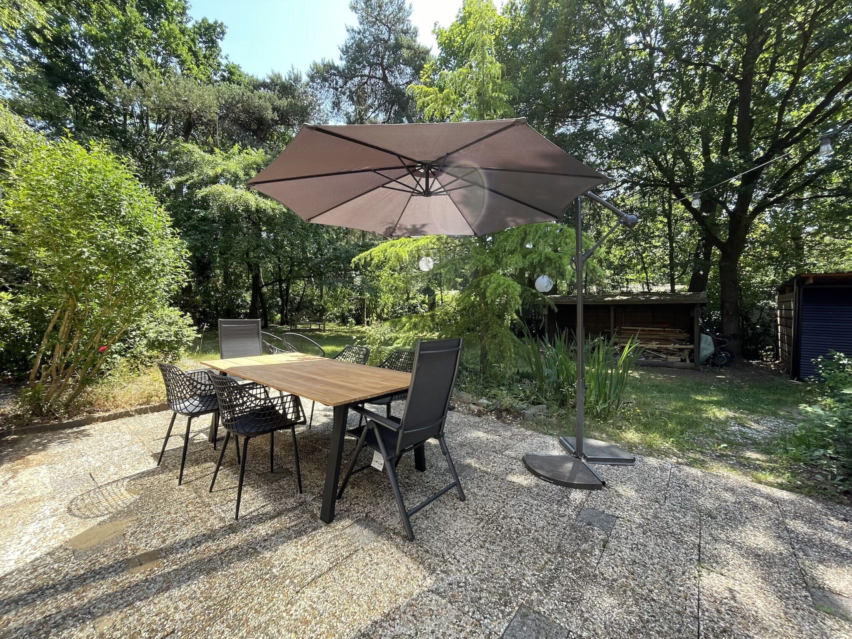 Un patio al aire libre con mesa y sillas de madera, un paraguas grande y suelo de grava.