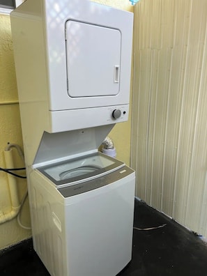 A compact, white washing machine with a front-loading door, placed against a yellow wall with a corrugated metal fence in the background. - "Stylish Studio Retreat - Outdoor Patio & kitchen (Miami)