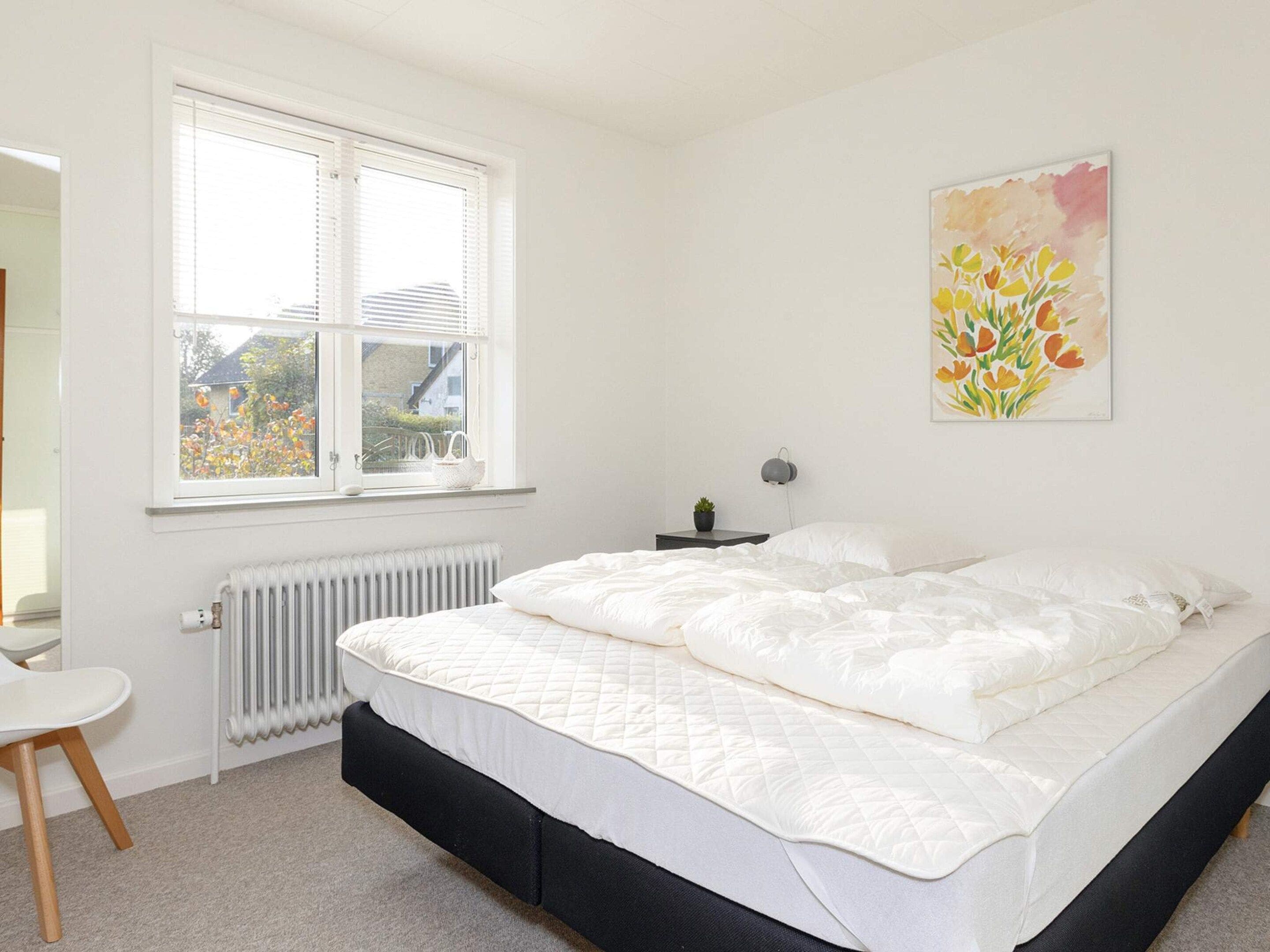 Quarto com cama, radiador, janela, mesa de cabeceira com lumin ária e um quadro na parede.