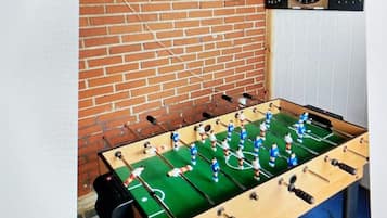 Uma mesa de futebol de mesa com jogadores de uniforme azul e branco, em frente a uma parede de tijolos com um alvo de dardos acima.