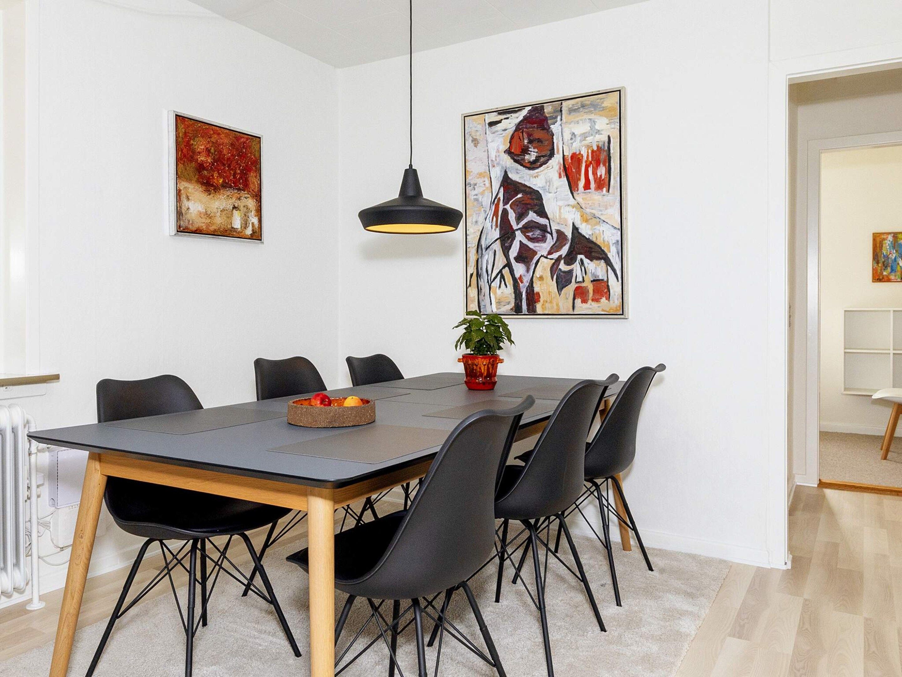 Un área de comedor con una mesa rectangular, sillas negras, una planta en maceta y dos cuadros abstractos en las paredes.