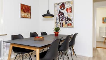 Un área de comedor con una mesa rectangular, sillas negras, una planta en maceta y dos cuadros abstractos en las paredes.