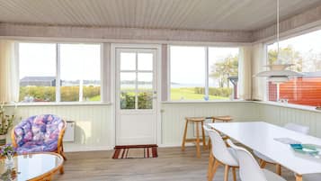Quarto com mesa de jantar, cadeiras, uma cadeira de vime, mesa de vidro e porta com vista para o exterior.