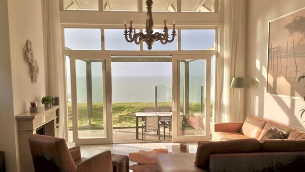 A spacious living room with a large sliding glass door leading to a patio with a view of the sea. There is a chandelier, a sofa, and a coffee table. - Dijkvilla on the Ijsselmeer (Makkum)