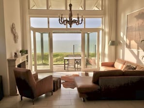 A spacious living room with a large sliding glass door leading to a patio with a view of the sea. There is a chandelier, a sofa, and a coffee table.