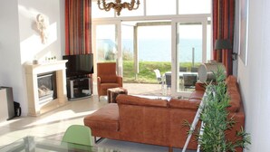 A spacious living room with a large sofa, a television, and a fireplace. There is a glass table and chairs, and a view of the sea through large windows. - Dijkvilla on the Ijsselmeer (Makkum)