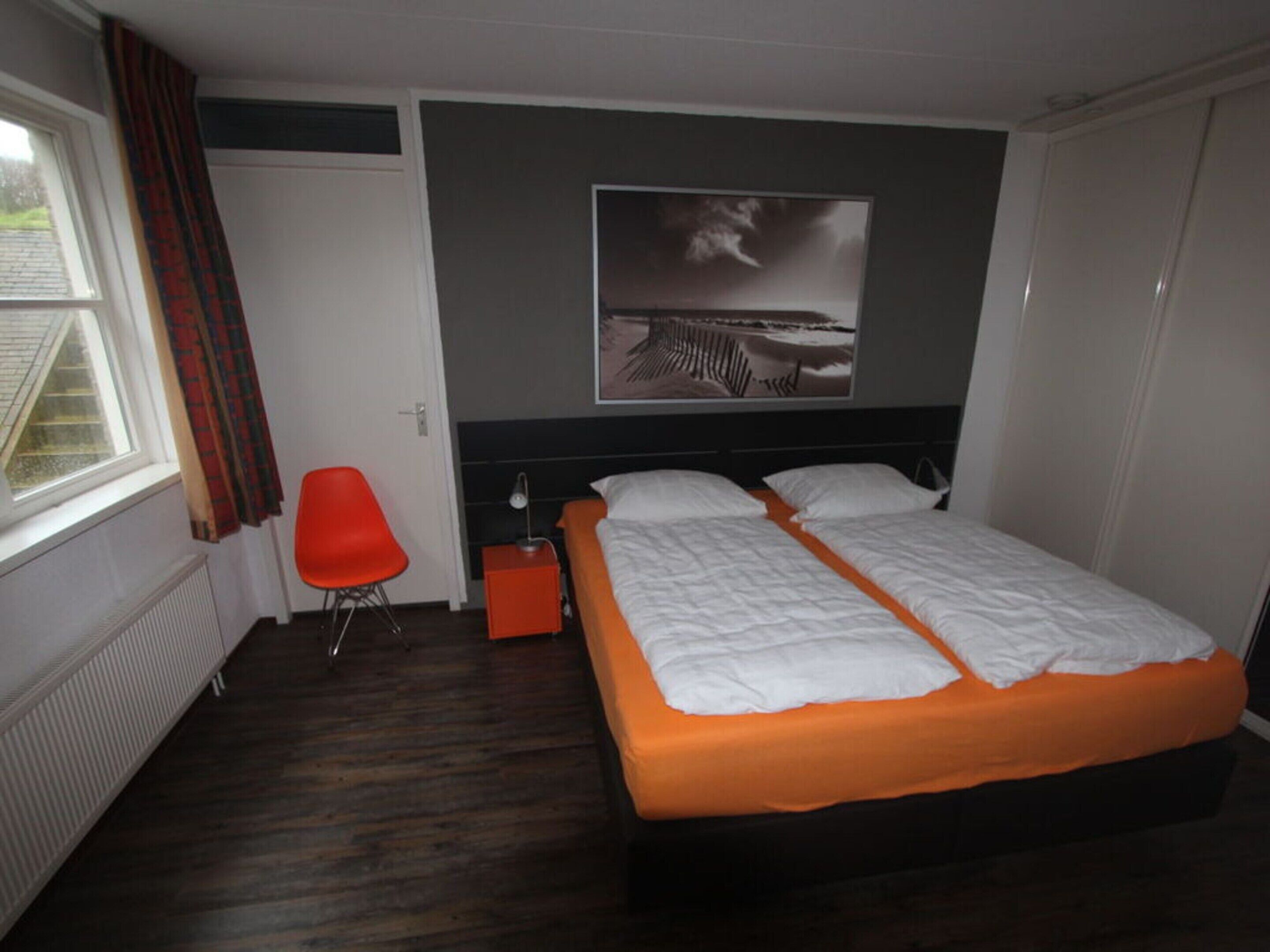 A bedroom with a double bed, a red chair, a nightstand, a window with curtains, and a wall-mounted picture.