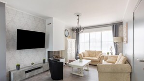 A modern living room with a flat-screen TV, a fireplace, a chandelier, and a large window. - Modern Villa w/ Private Yard (Middelkerke)