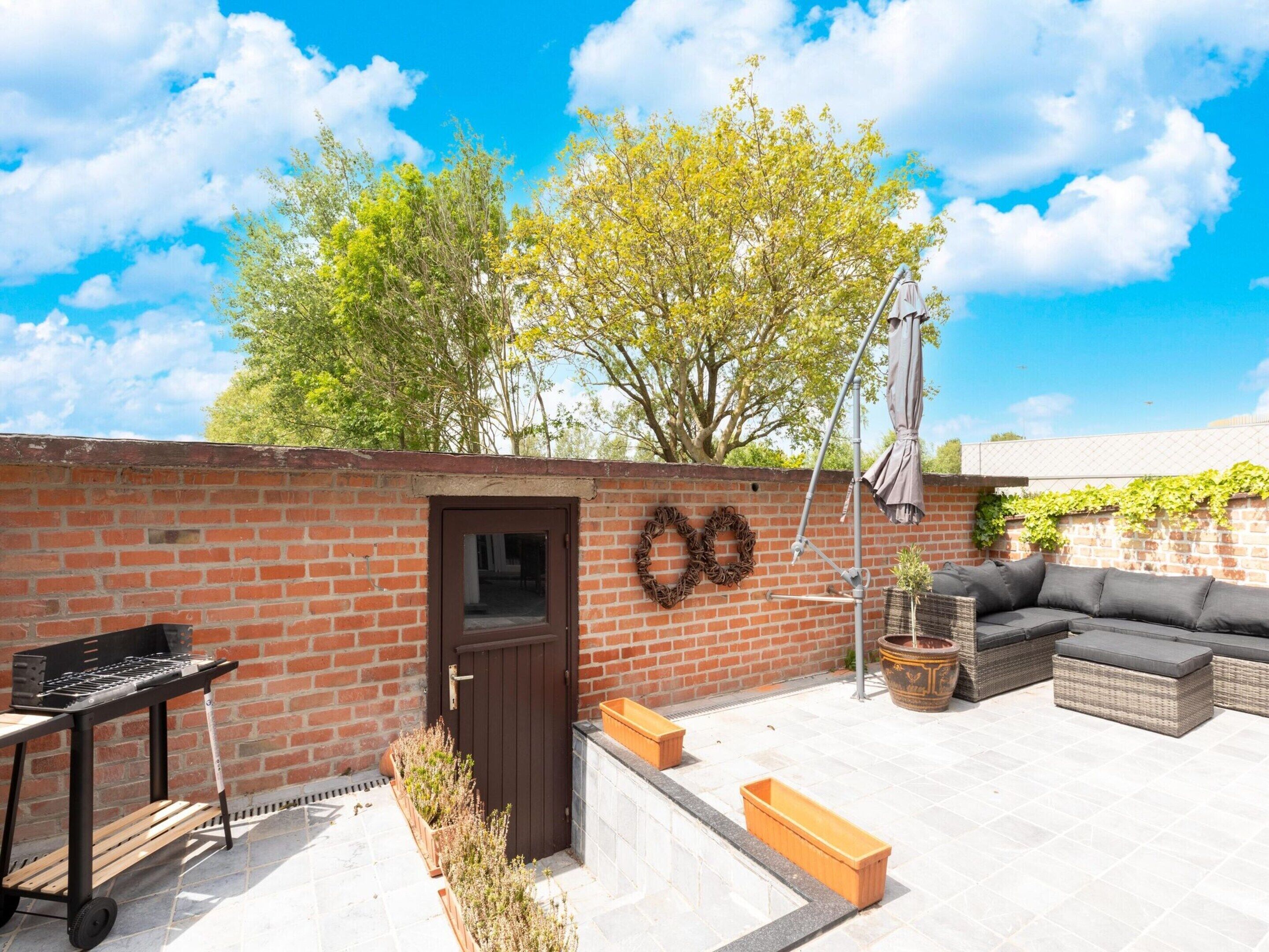 A patio with a brick wall, a grill, a door, a seating area with cushions, and a potted plant.