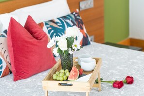 Ein Holztablett mit einer Vase voller weißer Blumen, einer Wassermelonenstange und grünen Trauben auf einem Bett mit gemustertem Bettbezug.