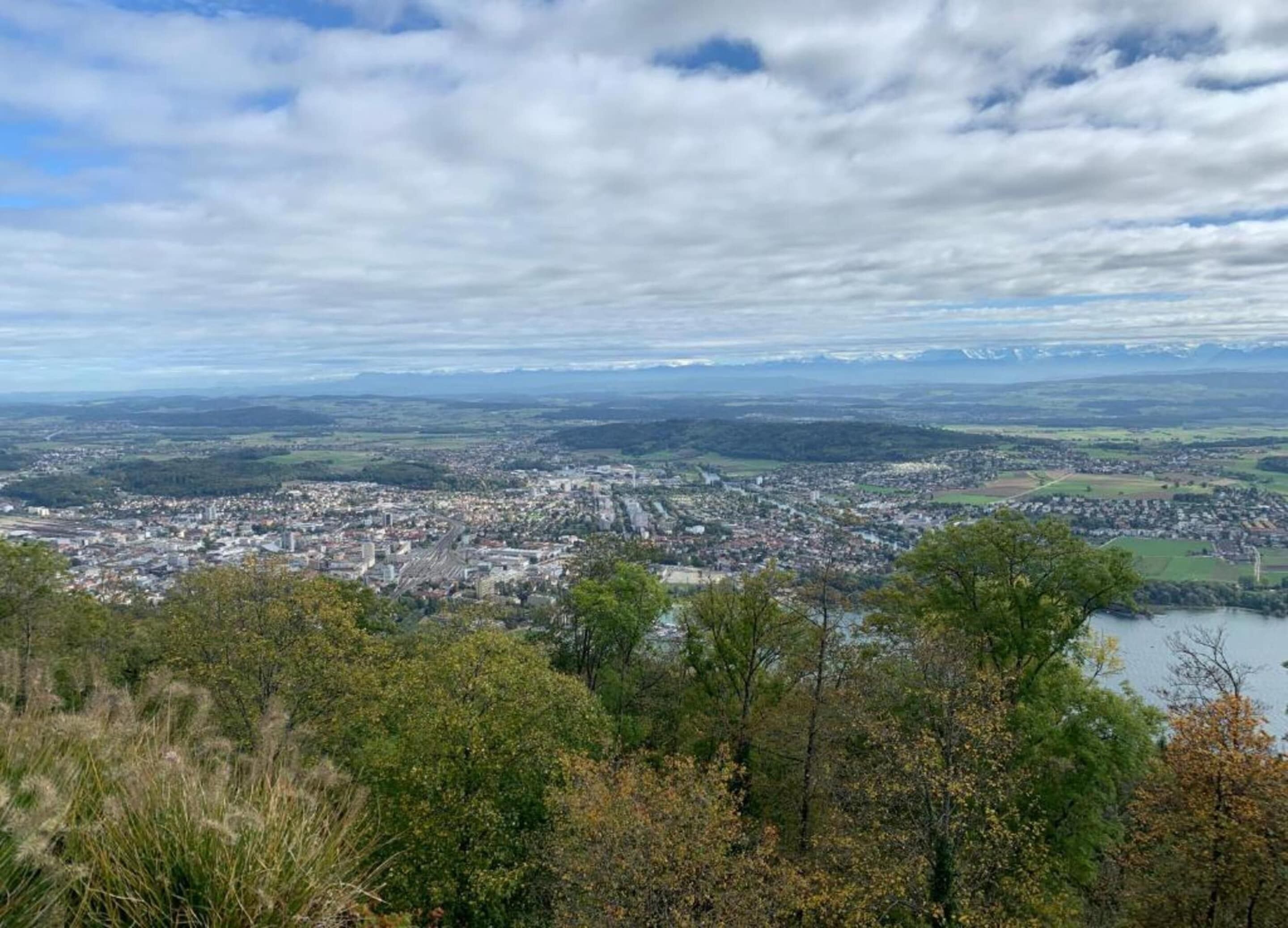 Une vue panoramique d’une ville, avec une rivière au premier plan, entourée de collines et d’arbres.